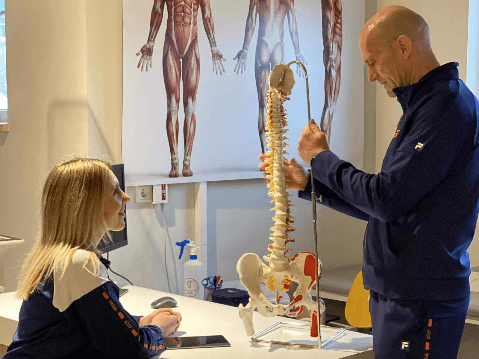 Eine Frau sitzt an einem Schreibtisch im INJOY Fitnessstudio Rudolstadt und lässt sich von einem Physiotherapeut an einem Wirbelsäulen-Skelett-Modell was erklären.