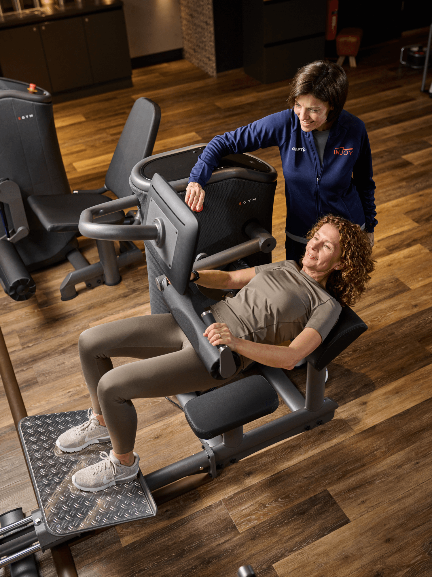 Ein weibliches INJOY Mitglied trainiert an einem EGYM Gerät und wird dabei von einer INJOY Trainerin betreut.