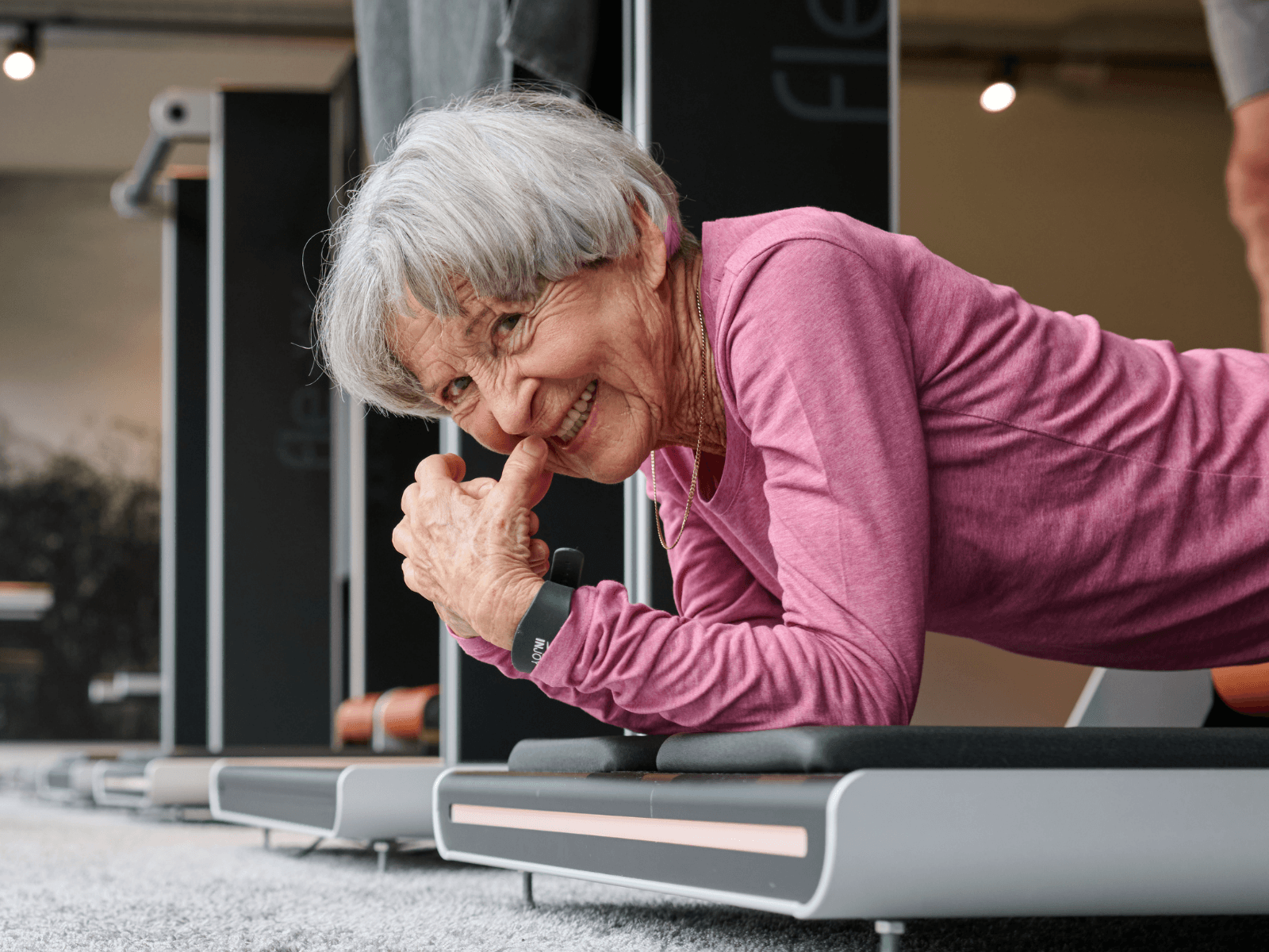 Seniorin in pinker Sportkleidung trainiert lachend an Gerät bei INJOY. Steht für gesundheitsorientierten Reha-Sport mit Spaß und persönlicher Betreuung.