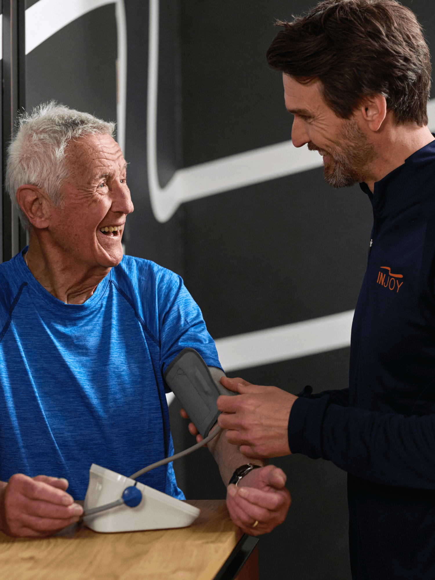 Ein INOJY Trainer misst bei einem älteren INJOY Mitglied im Fitnessstudio den Blutdruck.