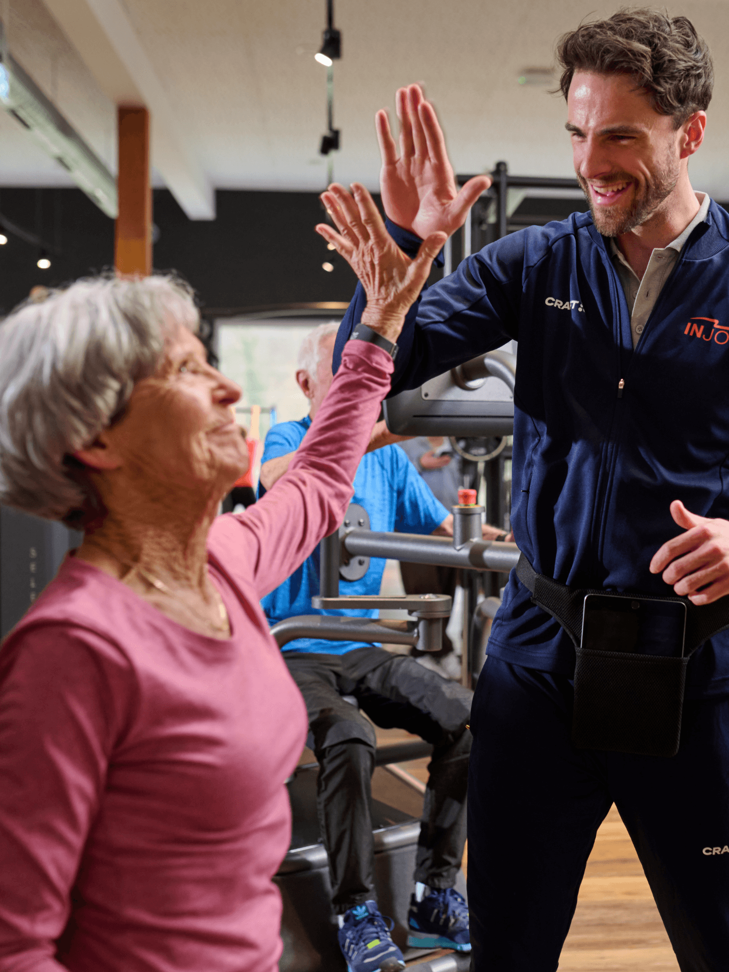 Ein älteres INJOY Mitglied sitzt an einem EGYM Gerät und klatscht dabei mit einem INJOY Trainer ab.