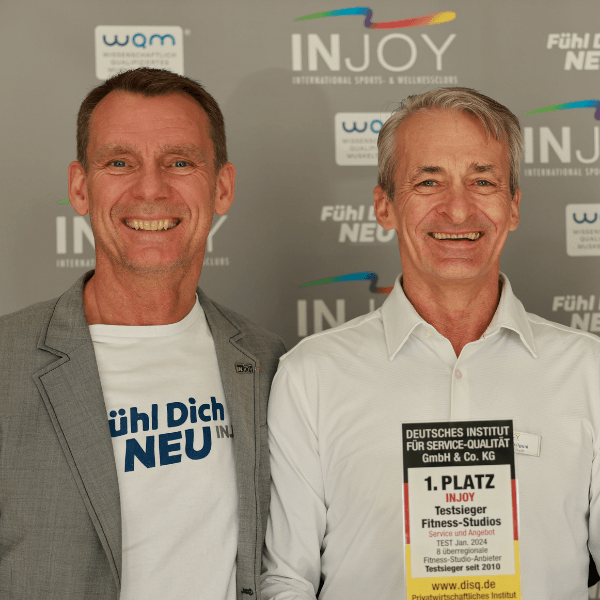 Thorsten Kielmann (GF INJOY QUALITY GmbH) und Herbert Weilguni (ehem. Betreiber INJOY Köln) posieren vor einer INJOY Fotowand und lächeln dabei in die Kamera.