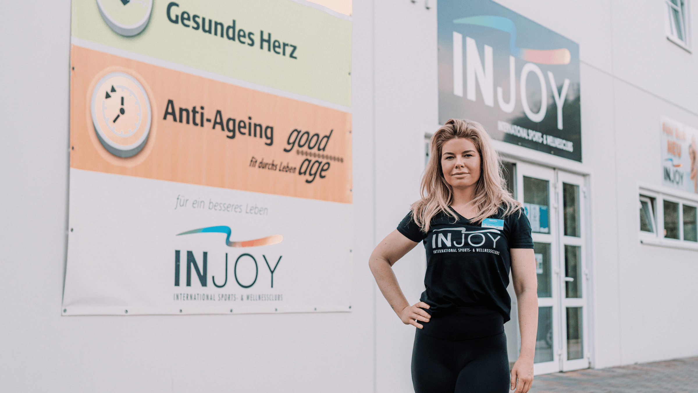 Eine INJOY Trainerin posiert stolz vor dem Eingang des INJOY Fitnessstudios in Waldkraiburg.