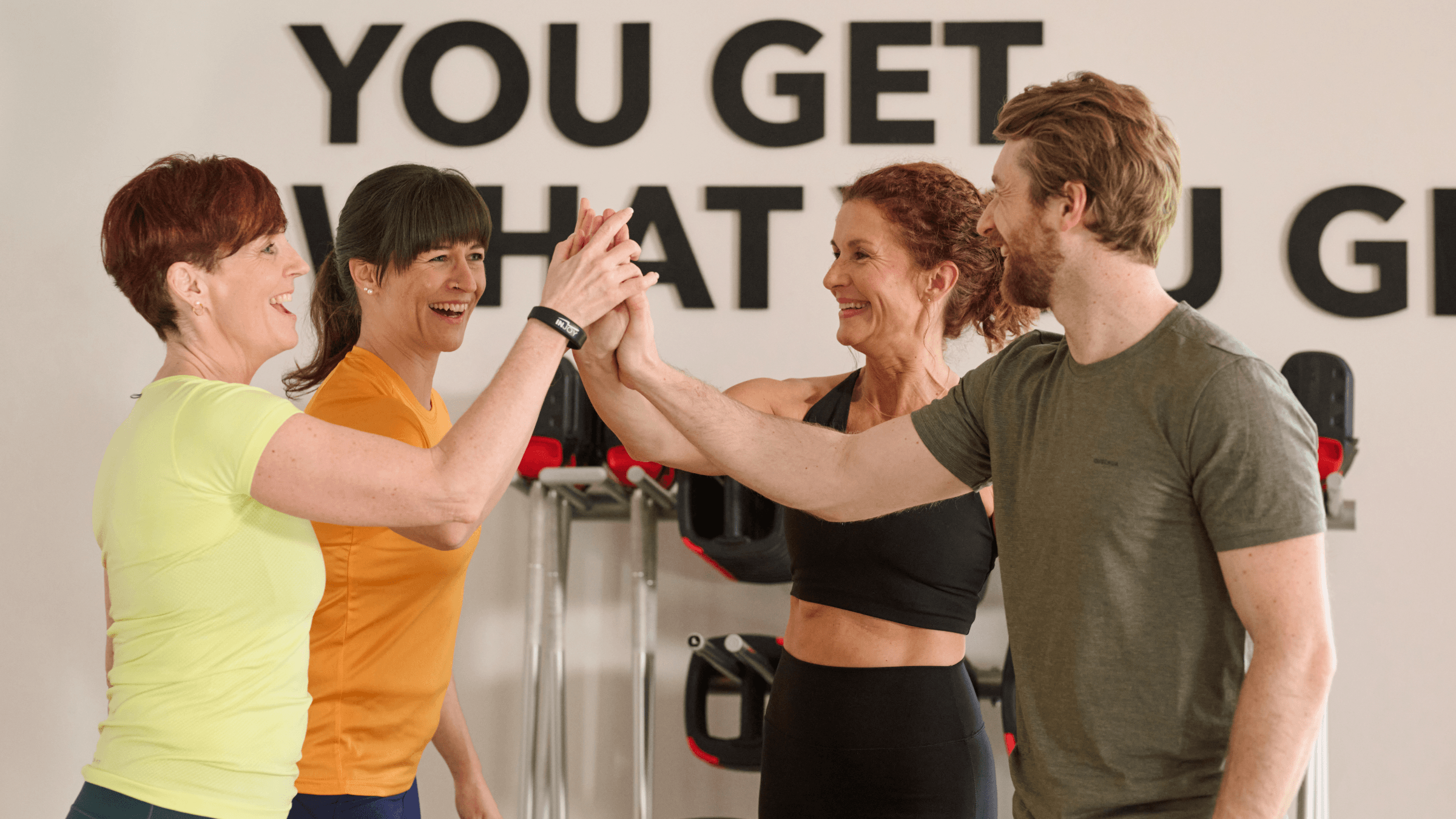 Eine Fitnessgruppe hat gerade einen Kurs beendet und klatscht gemeinsam ein. Sie stehen im Kursraum des Fitnessstudios.