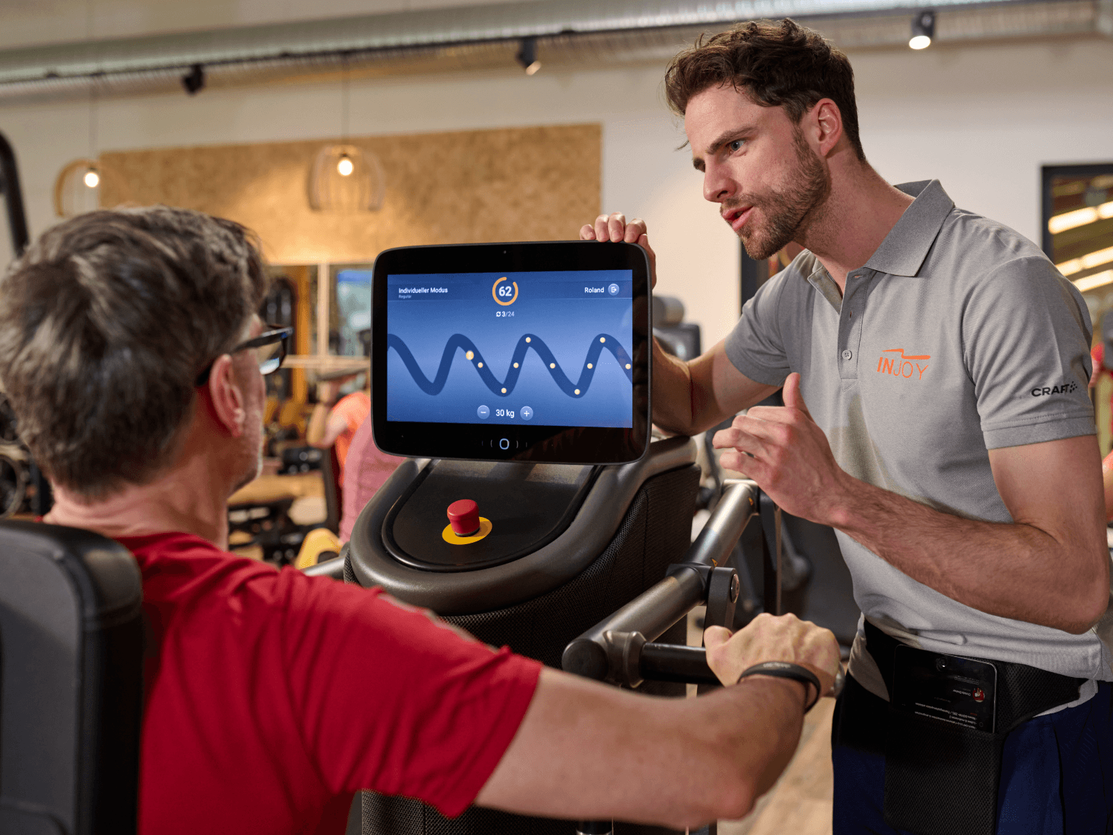 Ein junger INJOY Trainer gibt einem älteren Mitglied eine Einführung in ein EGYM Fitnessgerät.