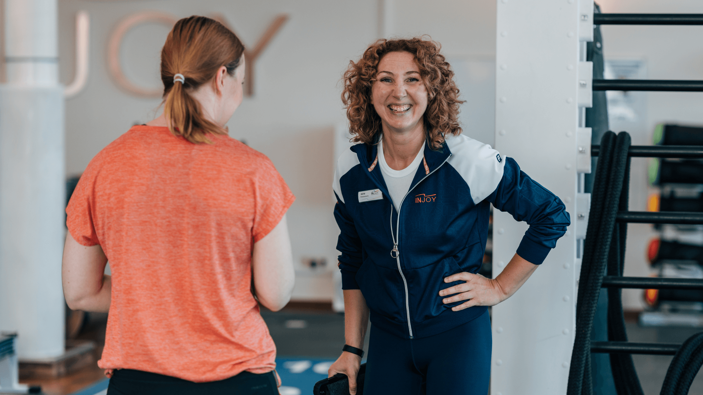 Eine lächelnde Frau steht im Hintergrund, während eine andere Frau vor ihr mit dem Rücken zur Kamera im INJOY Fitnessstudio Wolfsburg positioniert ist.
