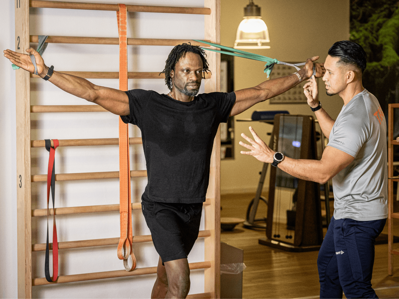 Ein Mitglied steht vor einer five-Sprossenwand im Fitnessstudio und dehnt mit zwei Therabändern die Brustmuskulatur. Er wird dabei von einem INJOY Trainer betreut.