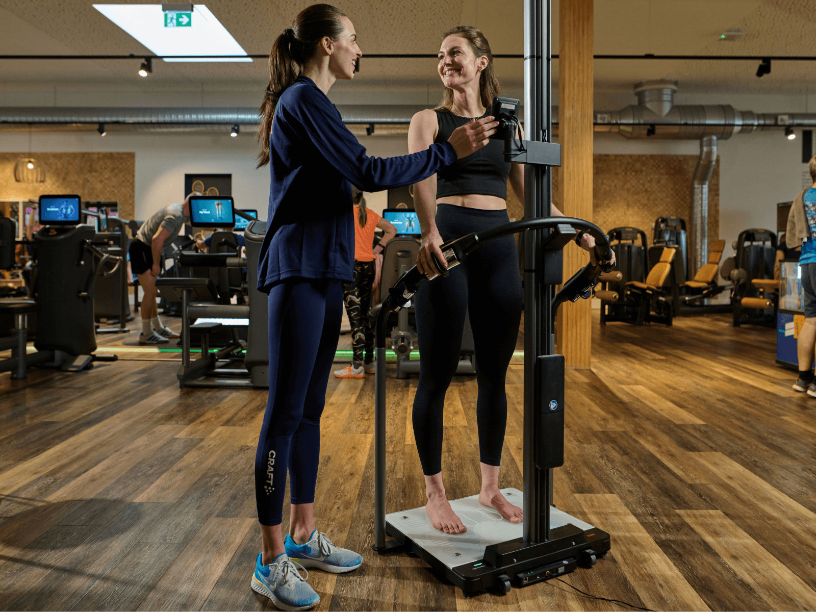 Ein weibliches Fitnessstudio-Mitglied steht auf einer SECA Waage und wird dabei von einer INJOY Trainerin unterstützt.