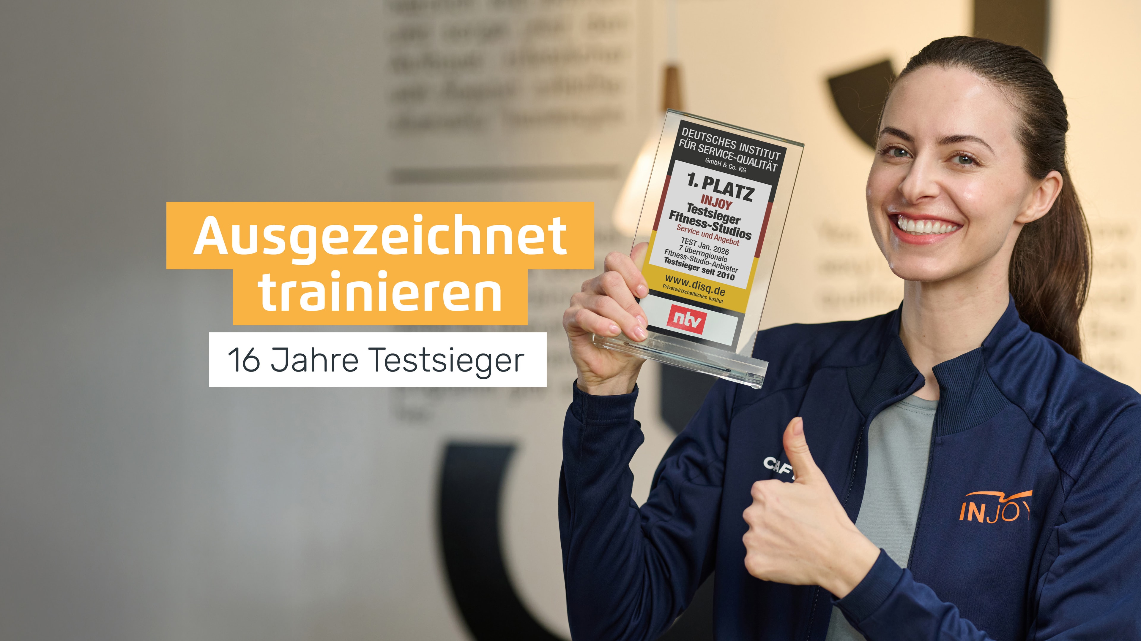 Eine fröhliche INJOY Trainerin hält den aktuellen INJOY Testsieger Award "Fitnessstudio 2026" in der Hand und streckt mit der anderen Hand einen Daumen nach oben.