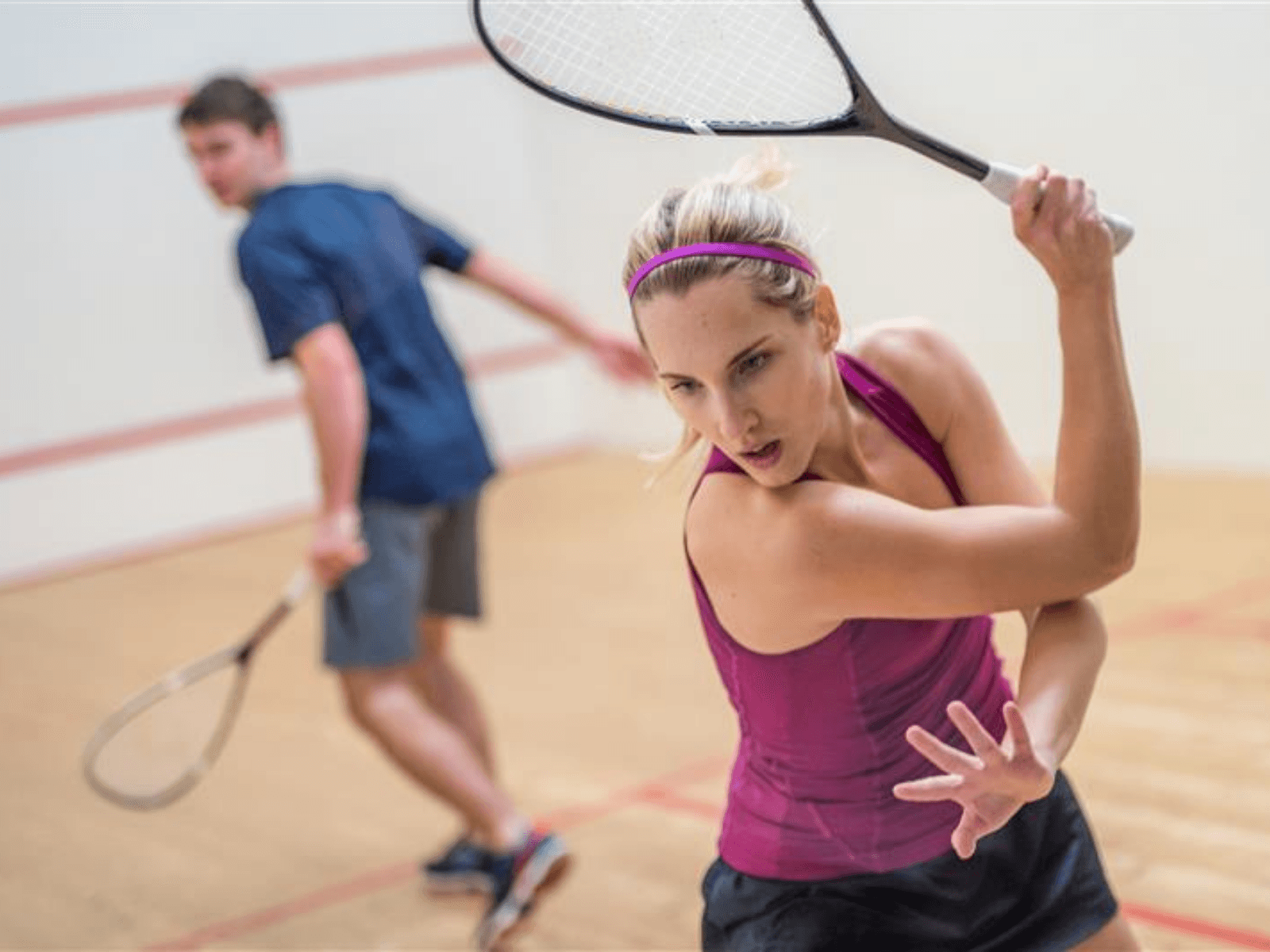 Ein INJOY Mitglied spielt Squash und verwendet ihre Rückhand, um den Ball im Spiel zu halten. Im Hintergrund sieht man ein männliches INJOY Mitglied, das gespannt auf den Ball schaut.