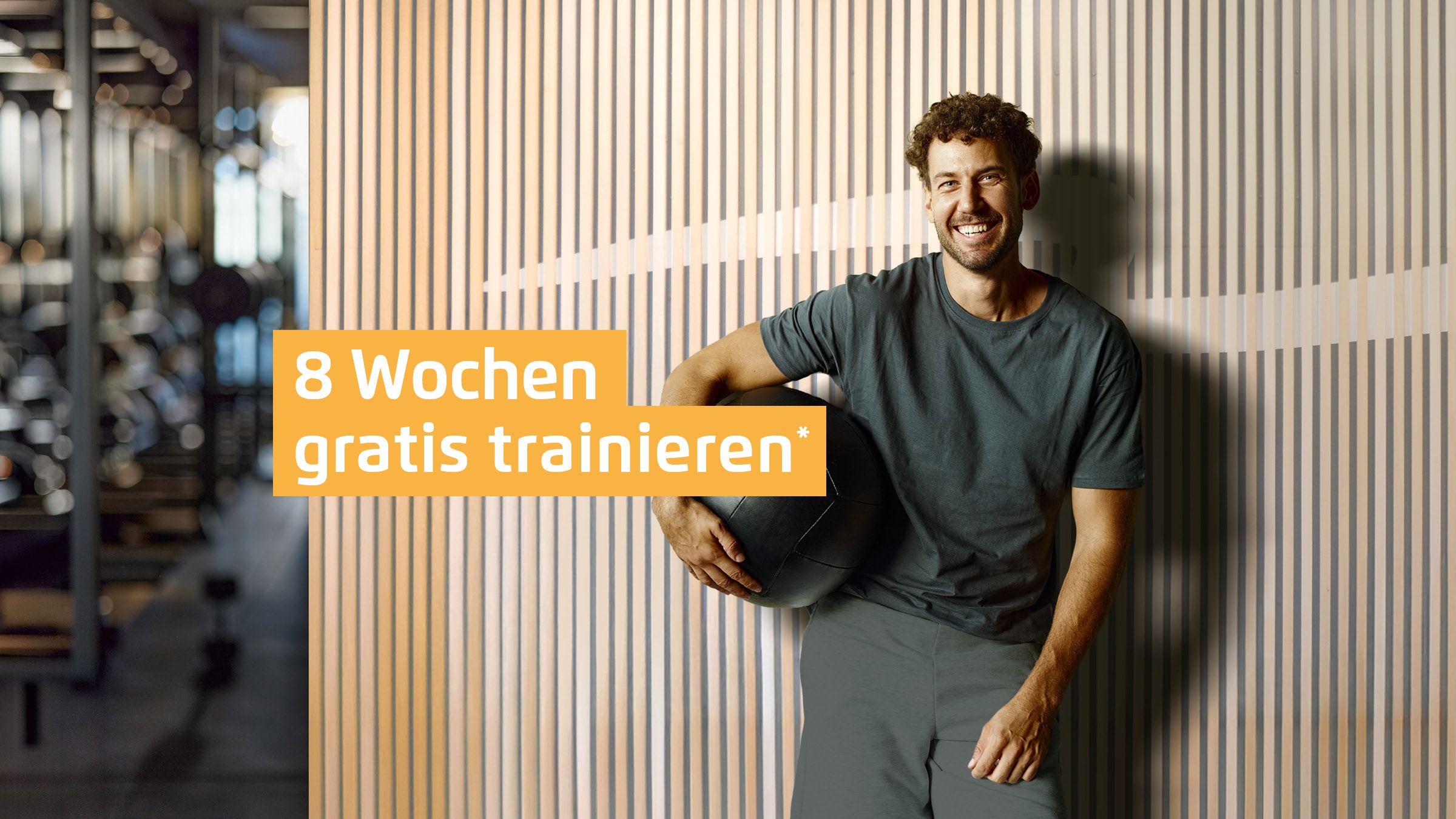 Ein männliches INJOY Mitglied posiert vor einer Holzwand mit INJOY Logo. Daneben ist der Slogan "8 Wochen gratis trainieren*" abgebildet".